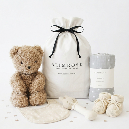 Neutral-toned deluxe gift hamper with a soft teddy bear, cream drawstring bag, gray rolled blanket, beige bib, soft toy, and