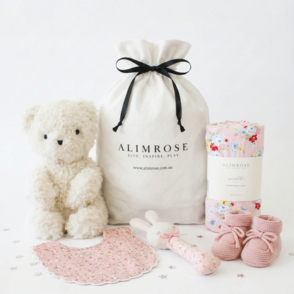 Pink-themed baby gift hamper with a plush teddy bear, floral blanket, rattle, booties, and white drawstring cotton bag with.