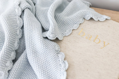 Powder blue knit baby blanket with moss stitch texture and scalloped edges draped over beige surface with "baby" text.