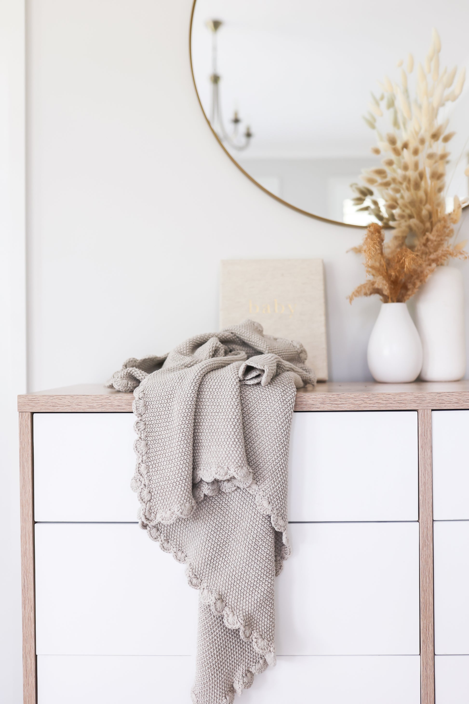 Latte-colored knit baby blanket with moss stitch pattern and scalloped edges draped over wooden dresser.