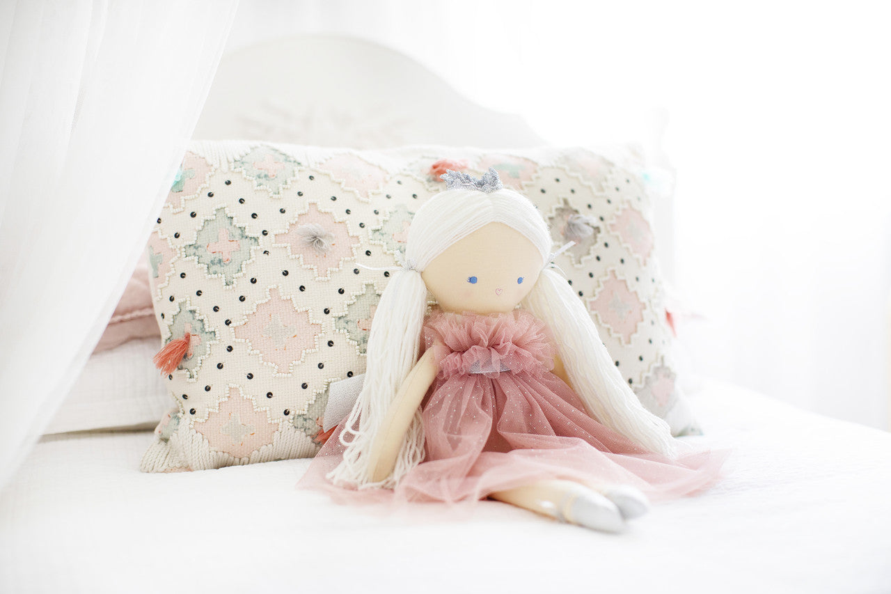 Penelope Princess doll in fluffy blush tulle frock with silver shoes, seated on bed with decorative pillow.