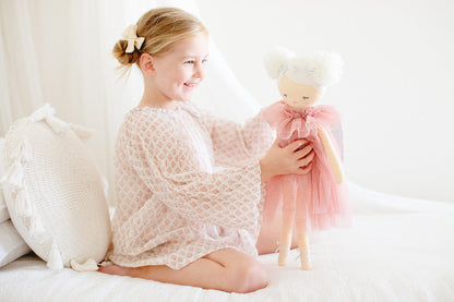 Smiling girl in a light dress holding the Ava Angel Doll, featuring a tulle dress and pom pom hairstyle.