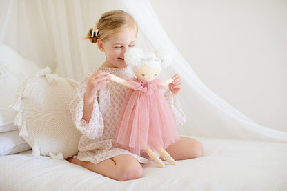 Girl sitting on a bed joyfully holding Ava Angel Doll, a 48cm pink tulle dress doll with pom pom hair and silver wings.